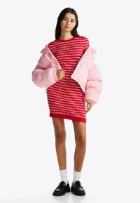 Rotes Kleid mit weißem Logo-Muster, kombiniert mit einer pastellrosa Steppjacke. Schwarze Slipper und weiße Kniestrümpfe runden das Outfit ab.