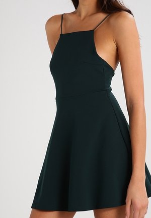 Ærmeløs mørkegrøn kjole med tynde stropper, firkantet udskæring, figursyet talje og udvidet nederdel båret af en stående person.