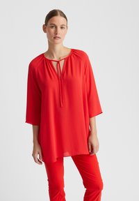 Rote, leichte Bluse mit Rundhalsausschnitt und Krawattendetail, drei Viertelarm, und lockerer Passform, kombiniert mit passenden roten Hosen.