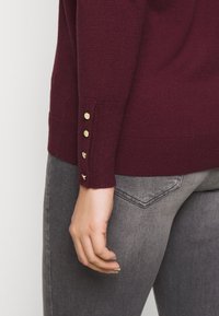 Pull en maille bordeaux avec un ourlet côtelé et des boutons dorés décoratifs sur le poignet. Associé à un jean gris avec un détail de poche arrière.