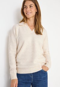Beige V-Ausschnitt-Pullover mit langen Ärmeln, gerippten Bündchen und Saum, weiche Textur, kombiniert mit blauen Jeans. Subtile horizontale Streifen.