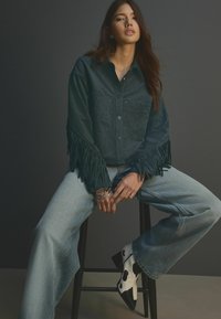 Dunkelgrünes Wildlederhemd mit Fransen, kombiniert mit hellblauen weit geschnittenen Jeans und schwarz-weißen Kuhflecken-High-Heels.