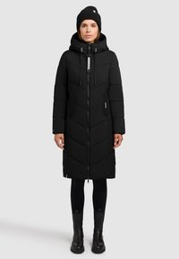 Abrigo largo negro aislante con cierre de cremallera, patrón en forma de chevrón, bolsillos laterales y capucha. Combinado con leggings negros y botas.