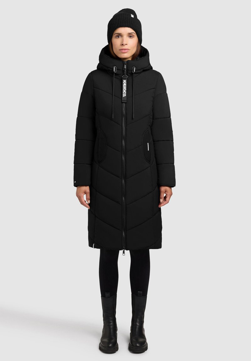 Abrigo largo negro aislante con cierre de cremallera, patrón en forma de chevrón, bolsillos laterales y capucha. Combinado con leggings negros y botas.