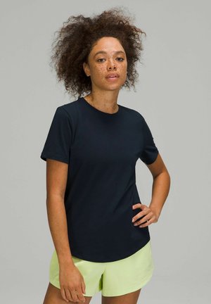Femme aux cheveux bouclés et aux taches de rousseur portant un t-shirt noir à manches courtes et un short vert clair, debout avec une main sur la hanche.