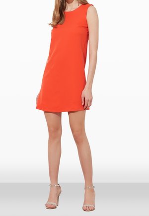 Woman wearing sleeveless, bright orange above-knee dress and silver high-heeled sandals, standing against a white background.