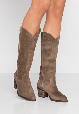 Jambes portant des bottes montantes en daim marron avec talons blocs empilés et bouts pointus, debout sur une surface blanche.