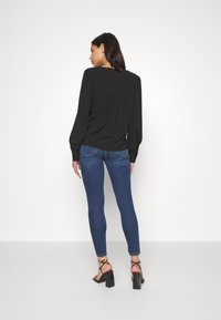 Svart långärmad blus med veck på baksidan, kombinerad med blå slim fit jeans och svarta klacksandaler. Slätt tyg med en figurnära design.