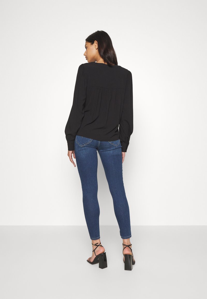 Blouse noire à manches longues avec des plis au dos, associée à un jean skinny bleu et des sandales à talons noires. Tissu lisse avec un design ajusté.