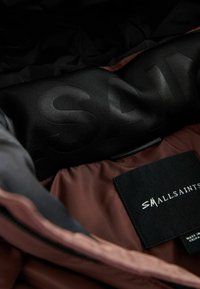 Brun quiltad jacka med svart foder, med en slät textur, logotryck och en skötsel etikett på högra sidan.