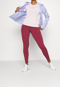 Lila lichtgewicht zip-up jack over een roze vochtafvoerende shirt, gecombineerd met bordeaux leggings en witte sportschoenen. Merkdetails zijn duidelijk zichtbaar.