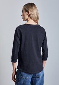 Laivansininen pitkähihaista paitaa, jossa on rento malli, raglan-hihat ja pyöreä kaula-aukko, yhdistettynä kuluneisiin sinisiin farkkuihin, joissa on koristeita.