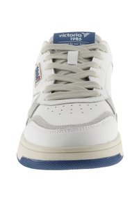 Zapatilla blanca con acentos grises, puntera redonda, suela texturizada y cordones prominentes. Presenta una etiqueta en la lengüeta azul con "victoria 1985."