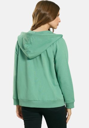 Kobieta o długich brązowych włosach, ubrana w zieloną, gładką bluzę z kapturem i czarne spodnie, stoi tyłem na jednolitym tle.