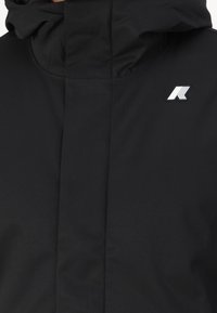 Chaqueta negra con un cuello alto, hecha de un material liso y resistente al agua. Presenta un pequeño logotipo blanco y un diseño aerodinámico.