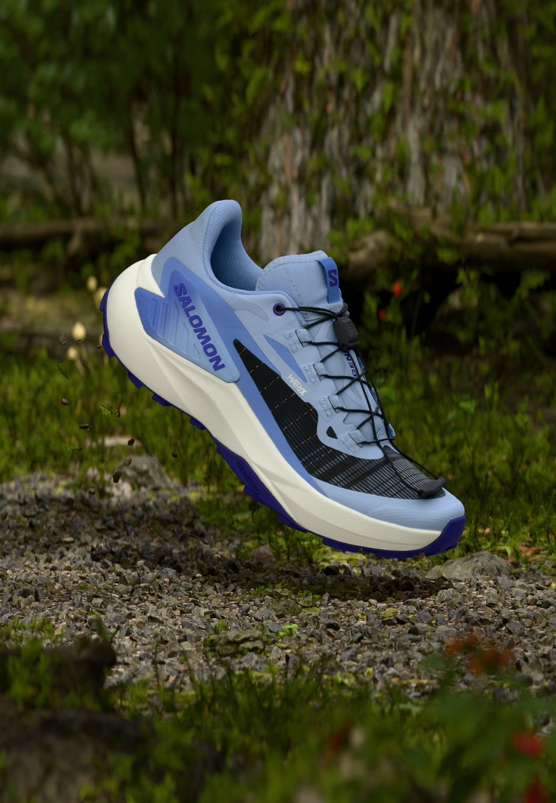 La zapatilla de trail running Salomon presenta una parte superior transpirable de color azul claro, diseño de malla negra, entresuela blanca y suela morada con agarre texturizado.