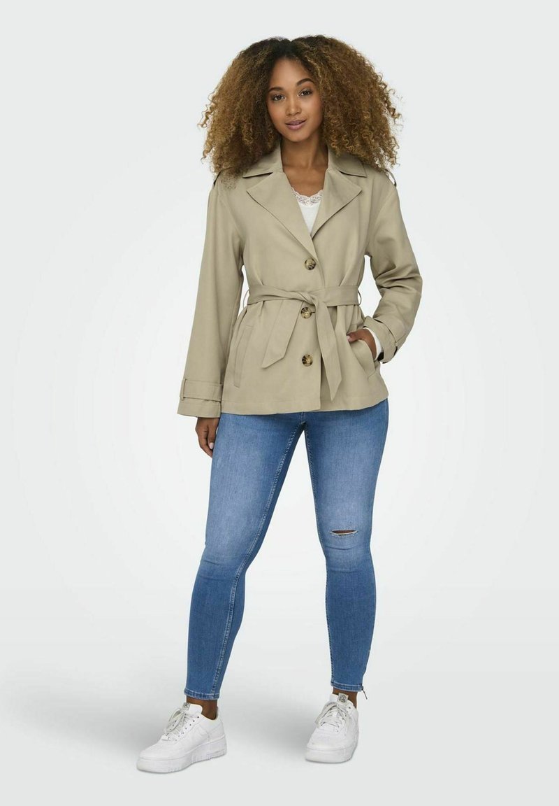 Manteau trench beige ceinturé avec de grands boutons, un col large et des manches longues, associé à un jean skinny bleu clair et des baskets blanches.