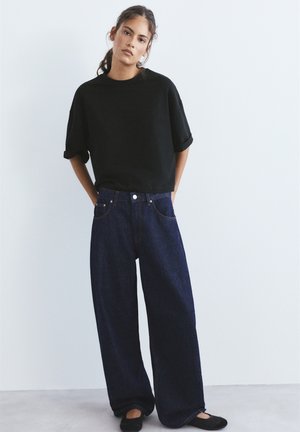 Junge Frau steht mit den Händen in den Taschen, trägt ein schwarzes, hochgekrempeltes Oberteil, weite dunkelblaue Jeans und schwarze Ballerinas vor einem einfarbigen Hintergrund.