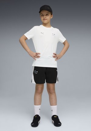 Barn står med hænderne på hoften iført en sort kasket, hvid Puma T-shirt, sorte shorts, hvide sokker og sorte Puma-sneakers foran en grå baggrund.