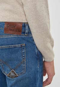 Persona che indossa jeans blu Roy Roger's e un maglione beige a maniche lunghe, mostrando la tasca posteriore e l'etichetta in pelle del marchio.