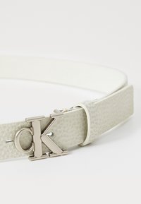 Calvin Klein diržas, pagamintas iš tekstūruotos šviesiai pilkos odos, su sidabro atspalvio "CK" logo sagtimi ir lygiu balto vidumi.