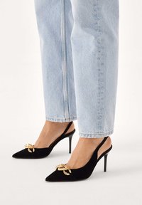 Černé semišové lodičky na podpatku s otevřenou patou, špičatou špičkou a zlatou ozdobnou mašlí. Mají štíhlý vysoký podpatek a design s otevřenou patou.