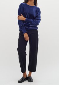 Blå sammet långärmad topp med rynkad axeldetalj, kombinerad med högmidjade svarta vida byxor och svarta loafers.