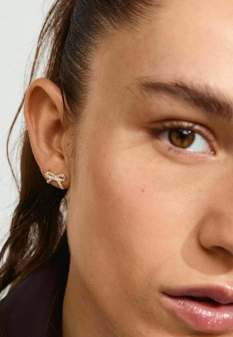 Zlato obarvani uhani v zasnovi z zavitjem, s prozornimi rhinestones, vgrajenimi vzdolž krivulj, pritrjeni na uho modela.