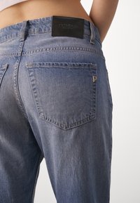 Los vaqueros de mezclilla azul claro presentan una cintura alta, un diseño de pierna recta y bolsillos traseros con costuras visibles. La pretina tiene una etiqueta de cuero negra.