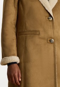 Cappotto in suede marrone con colletto in pelliccia shearling, caratterizzato da una sola fila di bottoni e tasche laterali. I polsini sono foderati in pelliccia shearling crema.