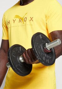 Černá kovová činka s texturovaným úchopem, se stříbrnou tyčí a červeným zámkem; drží ji osoba ve světlé žluté košili s logem.