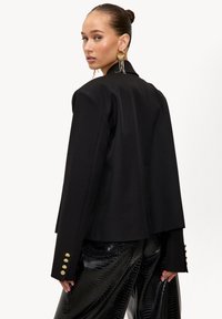 Blazer noir aux épaules structurées, fermeture à un bouton et détails en métal doré. Associé à un pantalon noir brillant à motifs.