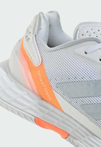 Harmaa urheilukengä, jossa on verkkomateriaali päällä, hopeiset yksityiskohdat, oranssi välikerros ja teksti "DEFIANT SPEED". Kengissä on pehmustettu kanta ja texturoitu pohja.