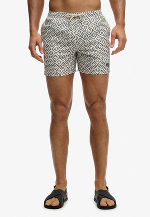 Męski model ubrany w beżowe kąpielówki z czarnym geometrycznym wzorem oraz czarne sandały na krzyżujące się paski, stojący na białym tle.