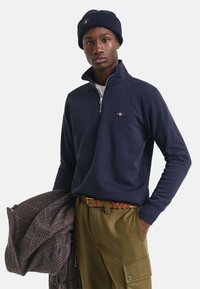 Marineblauwe zip-up sweatshirt met geribbelde kraag, gedragen met olijfgroenebroek en een gevlochten bruine riem. Houdt een gestreepte bruine jas vast.