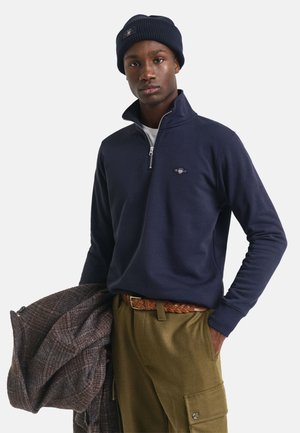 Marineblauwe zip-up sweatshirt met geribbelde kraag, gedragen met olijfgroenebroek en een gevlochten bruine riem. Houdt een gestreepte bruine jas vast.