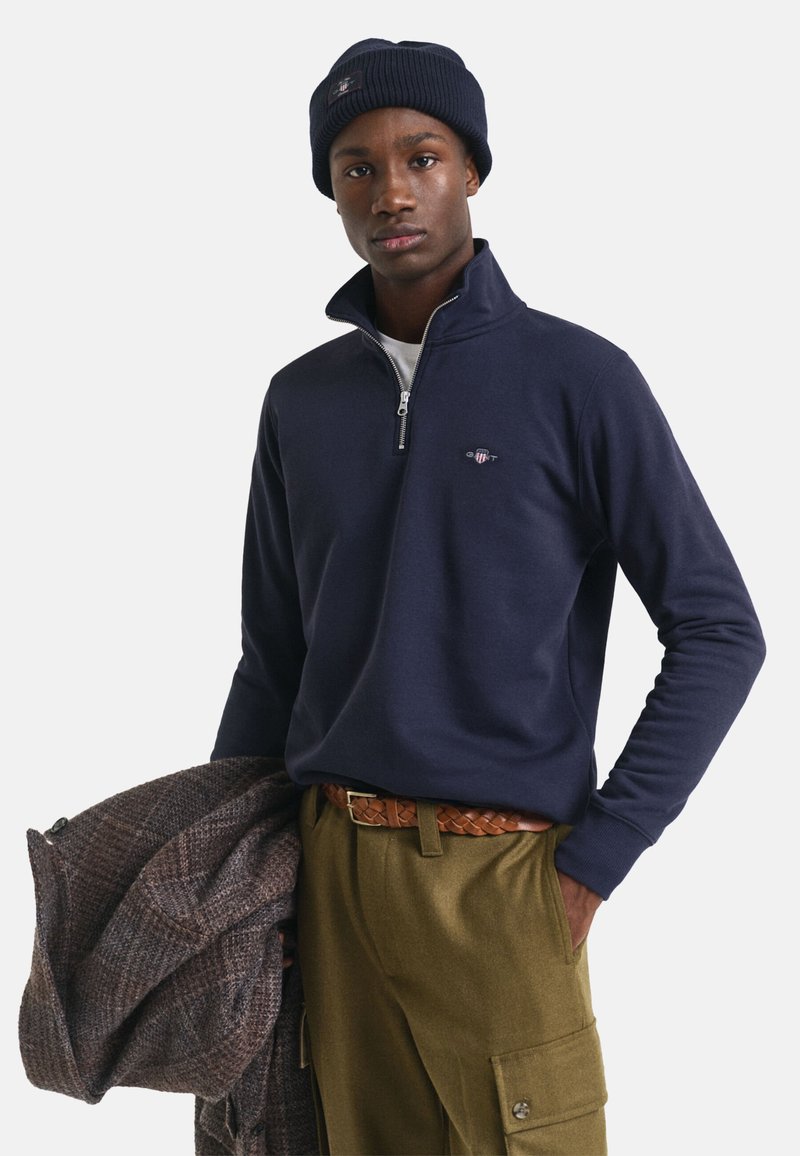 Marineblauwe zip-up sweatshirt met geribbelde kraag, gedragen met olijfgroenebroek en een gevlochten bruine riem. Houdt een gestreepte bruine jas vast.