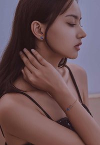 Pulsera de oro con acento azul, que presenta letras entrelazadas. Modelo con pendientes en forma de corazón a juego, piel suave y cabello oscuro.