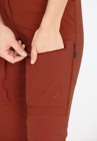 Ruosteenväriset vaellushousut, joissa on useita taskuja, vetoketjulliset sulkimet ja vahvistettu ompelujälki. Materiaali näyttää hengittävältä ja kestävältä.