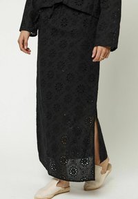 Zwarte maxi rok met bloemenperforaties, een zijsplit en een textuurstof. Gepaard met beige espadrilles.