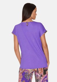Paarse t-shirt met korte mouwen en bloemenpatroon aan de randen. Heeft een kleine sleutelgaatje-opening aan de achterkant en is gemaakt van een zacht stof.