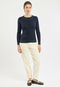 Gilet côtelé bleu marine avec des accents verts au col et à l'ourlet, doté d'une fermeture à boutons sur le devant. Associé à un pantalon en velours côtelé beige.