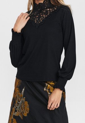 Kvinde iført en sort bluse med lange ærmer og blondedetalje ved halsen samt en sort nederdel med store brune blomsterprint.