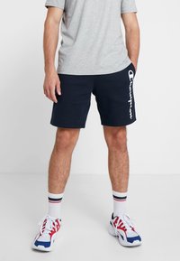 Marinblå shorts med vitt "Champion"-logotyp på sidan, kombinerat med en grå t-shirt, vita strumpor med röda och blå ränder, och flerfärgade sneakers.