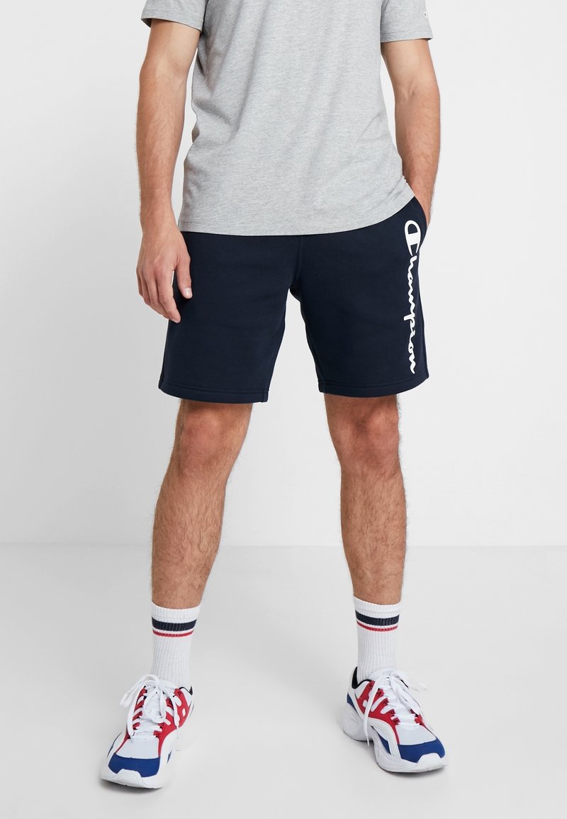 Marinblå shorts med vitt "Champion"-logotyp på sidan, kombinerat med en grå t-shirt, vita strumpor med röda och blå ränder, och flerfärgade sneakers.