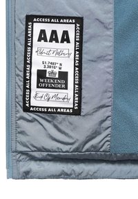 Zwart-witte stoffen patch op een blauwe jas met de tekst: "AAA Toegang Tot Alle Gebieden," coördinaten, "Weekend Overtreder," en handgeschreven zinnen.