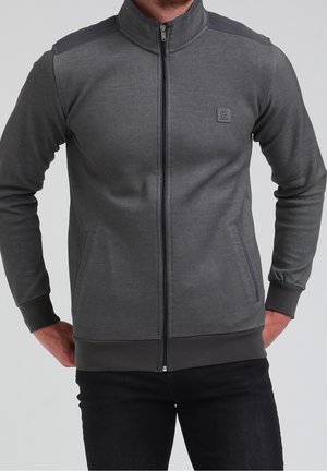 Mann trägt eine dunkelgraue Zip-Up-Jacke mit Taschen und einem kleinen quadratischen Logo auf der linken Brust, kombiniert mit schwarzen Hosen.