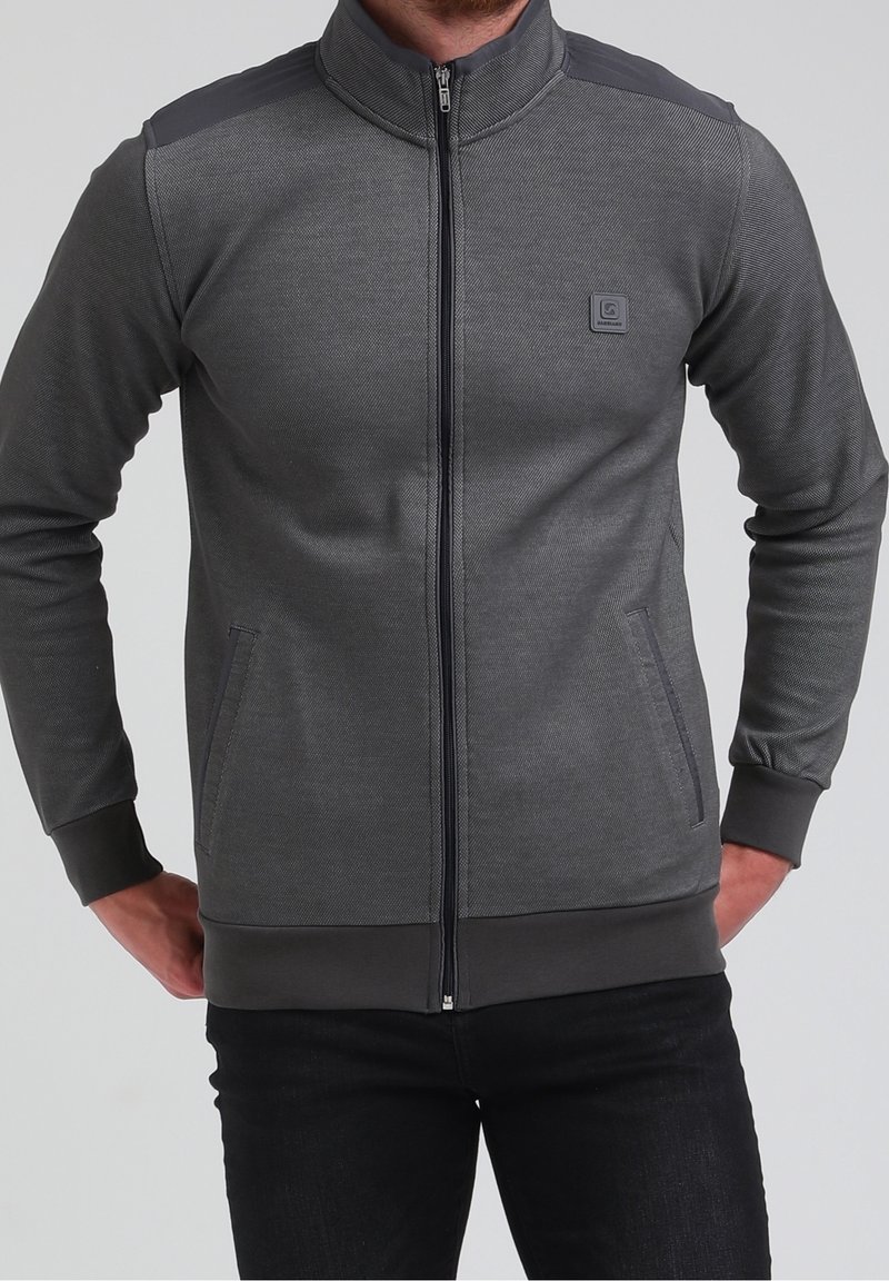 Homme portant une veste zippée gris foncé avec des poches et un petit logo carré sur la poitrine gauche, associée à un pantalon noir.