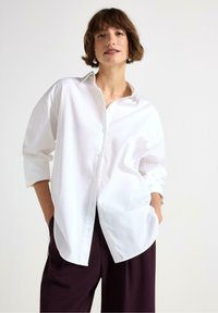 Kvinde med kort brunt hår iført oversized hvid skjorte med knapper og løse mørke bukser, stående med hænderne i lommerne mod en hvid baggrund.