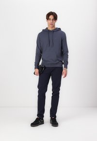 Felpa blu navy con tasca frontale e coulisse regolabili, abbinata a pantaloni scuri e sneakers nere, su uno sfondo bianco.